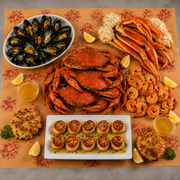 A seafood spread arranged on a patterned orange table mat, featuring cooked mussels, lobster legs, shrimp, scallops on a white platter, crab meat, two crouches, lemon wedges, and small cups of beer.