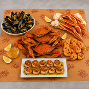 A seafood spread arranged on a patterned orange tablecloth, featuring cooked mussels in a white bowl, large red-orange crabs, lobster legs, shrimp, scallops on a white platter, lemon wedges, and a small glass of amber sauce on a gray surface.